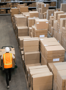 Worker inspecting large stacks of cardboard boxes in a warehouse.